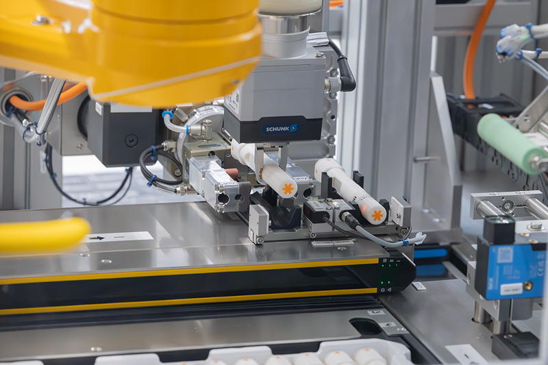 Stabiles automatisiertes Handling selbst bei komplexen Produktgeometrien: Während der Greifer einen etikettieren Autoinjektor zurück ins Tray legt, bewegt die Transport- und Rotationseinheit bereits ein neues Produkt Richtung Spendekante des Etikettierers.